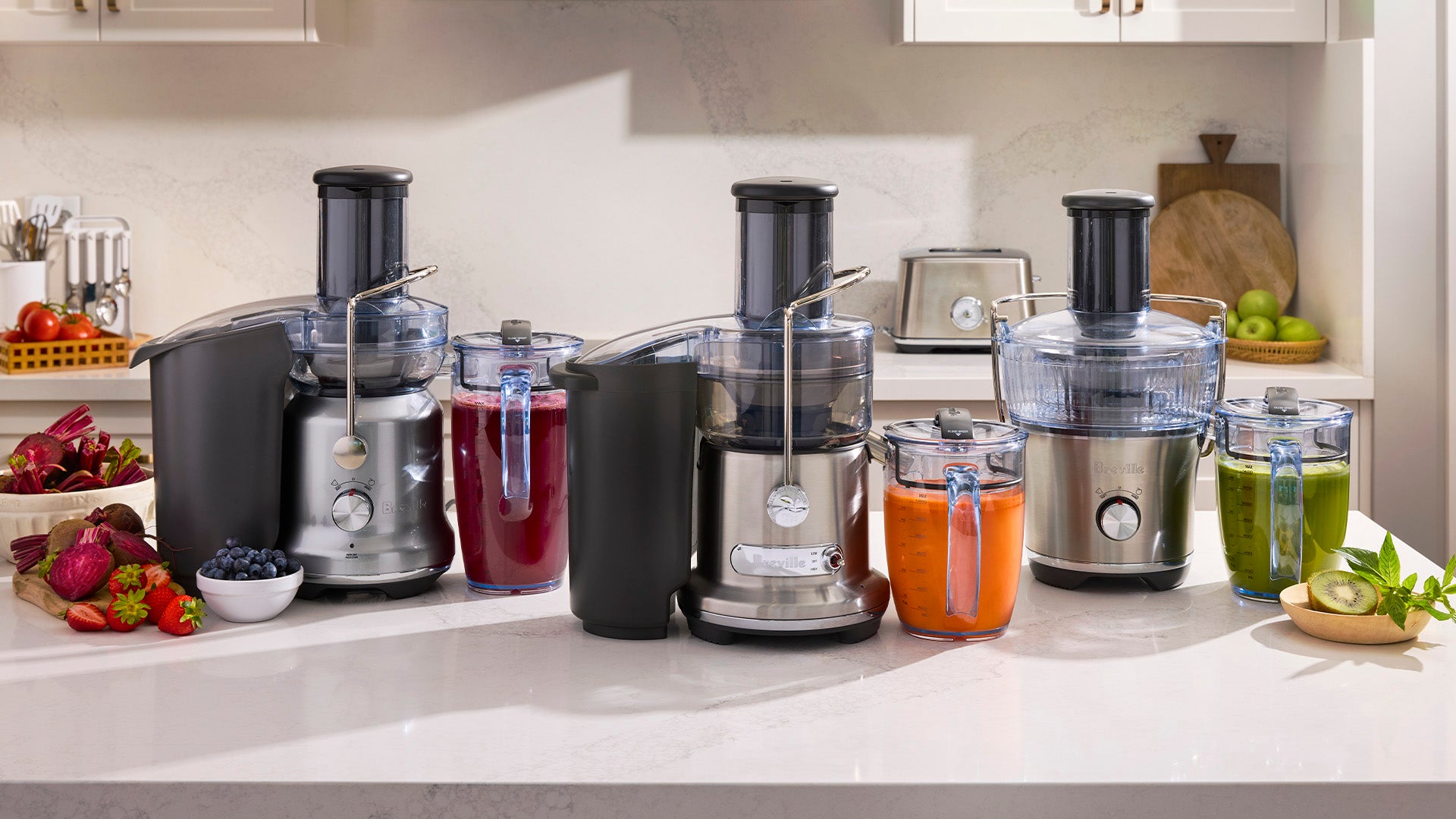 Juicers displayed on a kitchen countertop with glasses of fresh beetroot, carrot and green juice, surrounded by fresh fruit and vegetables.