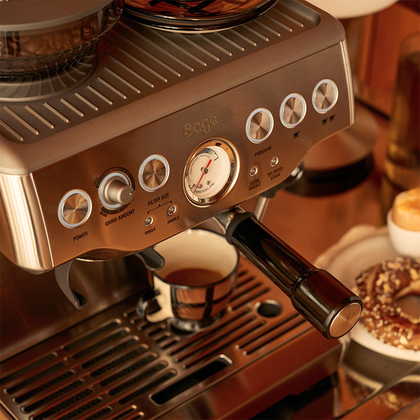 Sage Barista Express espresso machine on wooden sideboard, beans in hopper, espresso flowing into cup with cups and lamp arranged alongside