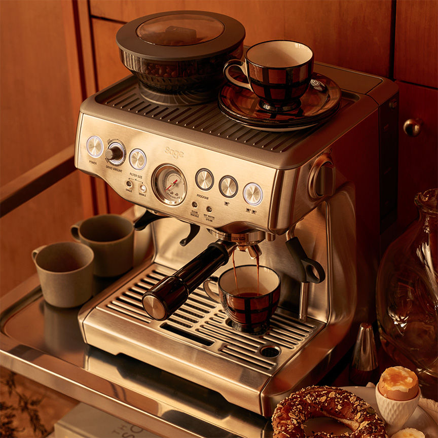 Sage Barista Express espresso machine on wooden sideboard, beans in hopper, espresso flowing into cup with cups and lamp arranged alongside