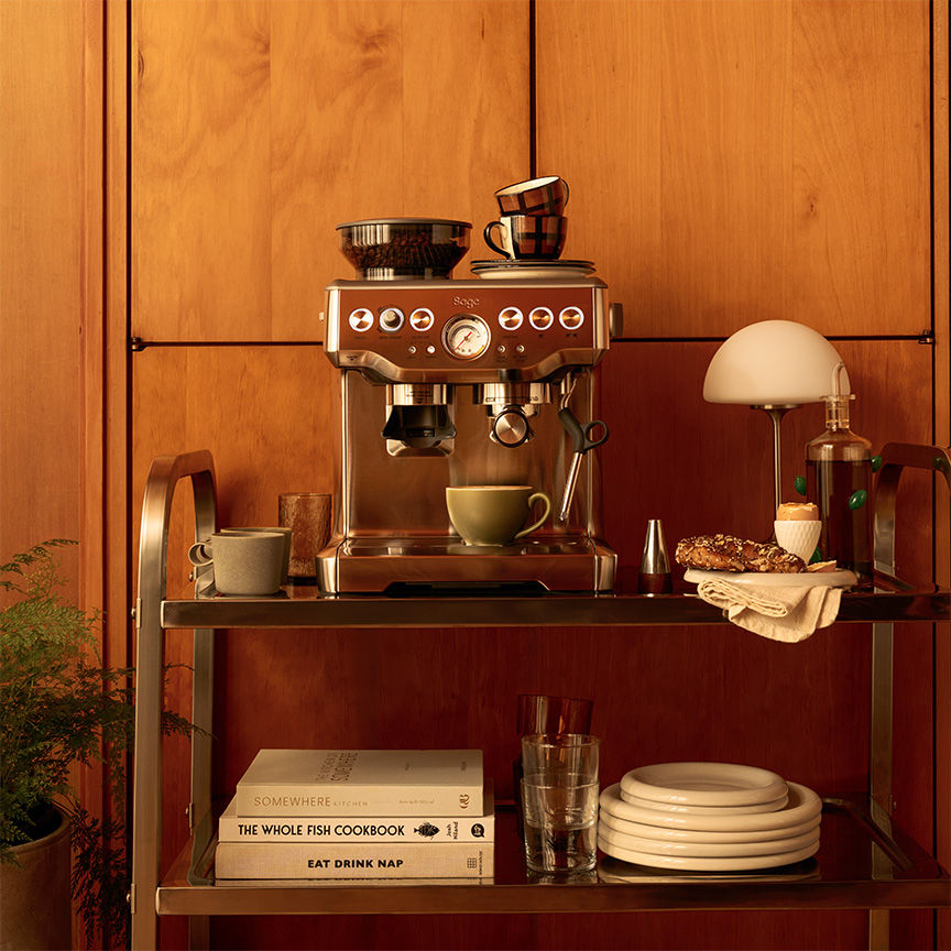 Sage Barista Express espresso machine on wooden sideboard, beans in hopper, espresso flowing into cup with cups and lamp arranged alongside